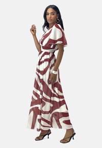 Robe maxi en blanc et bordeaux avec un motif abstrait ondulé, des manches courtes flottantes et une taille smockée. Elle est accompagnée de talons noirs ouverts.