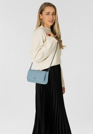 Vrouw draagt een roomkleurige trui en een zwarte geplooide rok en houdt een lichtblauw schoudertasje met kettingriem over haar schouder.