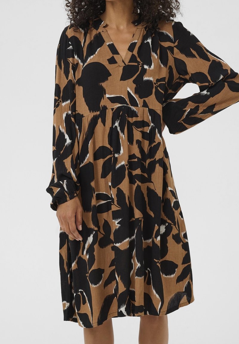 Femme portant une robe marron à manches longues avec de grands motifs floraux abstraits noirs et blancs, main reposant sur la hanche et pose détendue.