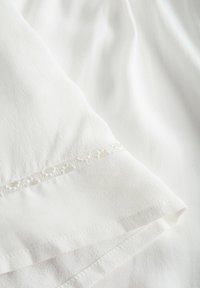 Tessuto bianco con una superficie testurizzata. Presenta un sottile bordo in pizzo con piccoli ritagli lungo il margine. Drape morbido e finitura liscia.