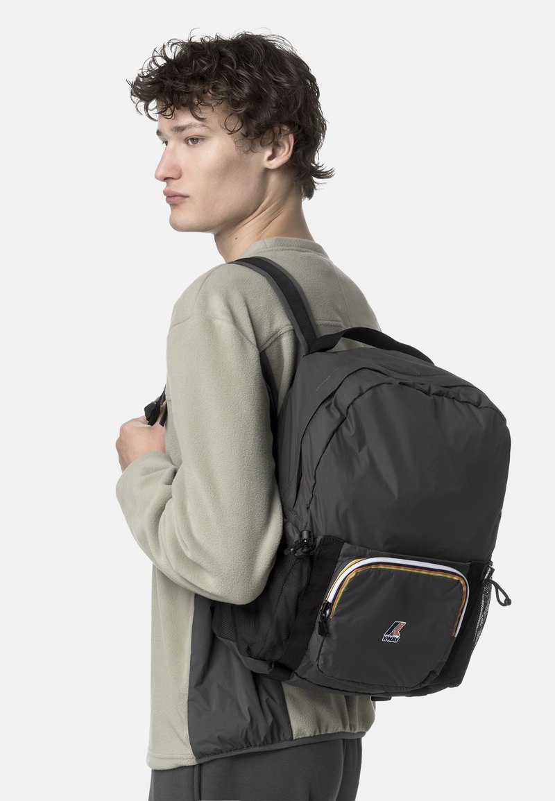 Zaino imbottito nero con tasca frontale con zip, dettagli arancioni e bianchi e tasche laterali in rete. Indossato su una spalla.