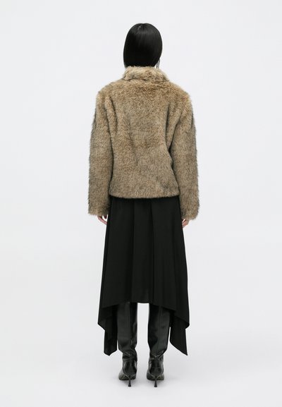 Ψεύτικο καμηλό παλτό με μαλακή υφή, φορεμένο πάνω από μια μαύρη ασυμμετρία φούστα και εφαρμοστά μαύρα παντελόνια, αναδεικνύοντας έναν στρωτό σχεδιασμό.