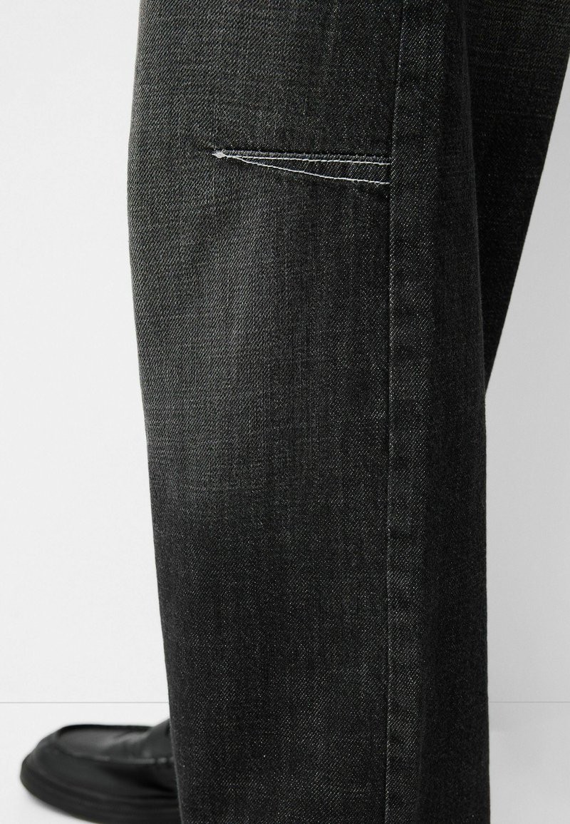 Pantalons en denim noir avec une finition légèrement texturée, dotés d'une poche cousue et d'une coupe droite, associés à des chaussures noires.