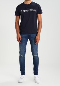 T-shirt en coton navy avec le logo blanc "Calvin Klein", associé à un jean skinny bleu déchiré et des baskets blanches. Look épuré et décontracté.