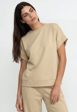 Naine pikkade tumedate juustega, kes kannab beežikat lühikeste varrukatega toppi ja sobivaid pükse, poseerib valgel taustal ja vaatab veidi kõrvale.