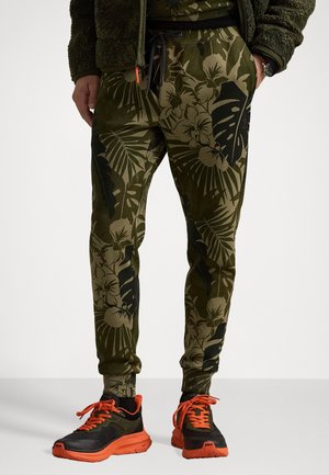 Groene bloemen sweatpants met een gedrukt ontwerp, een elastische tailleband met trekkoord en boordjes aan de enkels. Gemaakt in combinatie met zwarte en oranje sportschoenen.