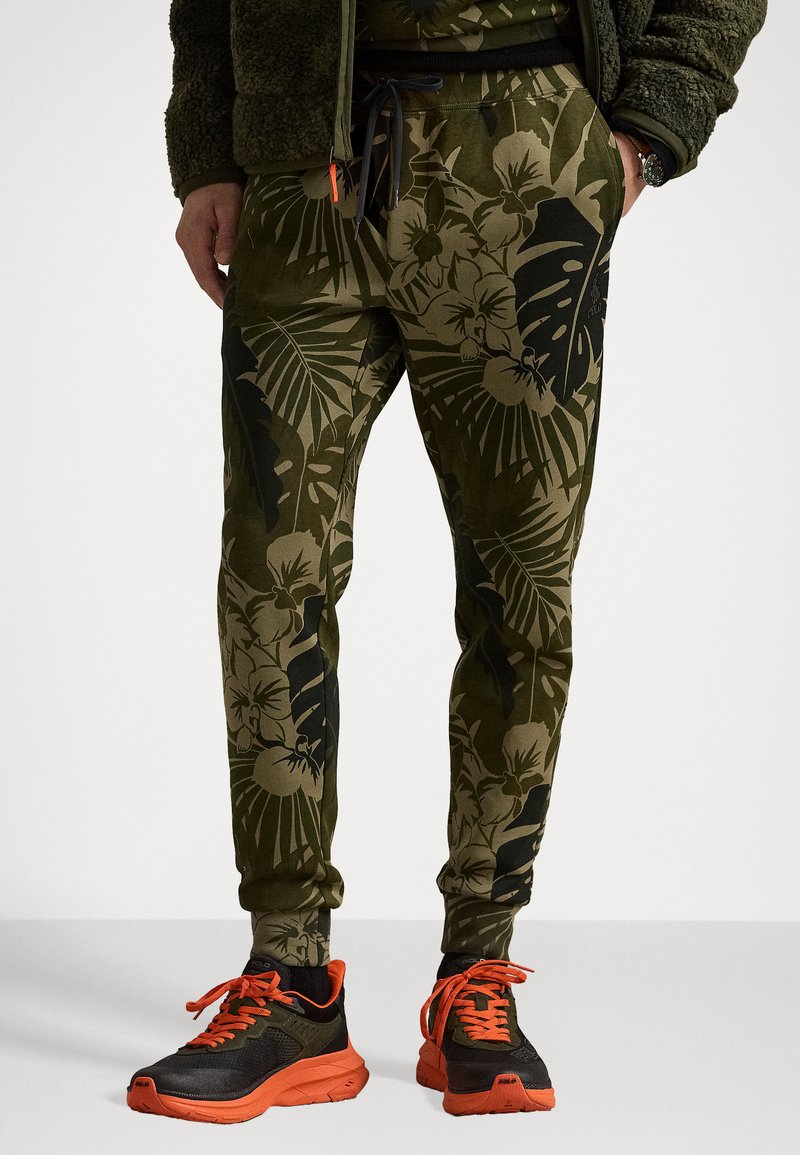 Pantalones de chándal verdes con un diseño floral, cintura elástica con cordón y tobillos ajustados. Combinados con zapatos deportivos negros y naranjas.