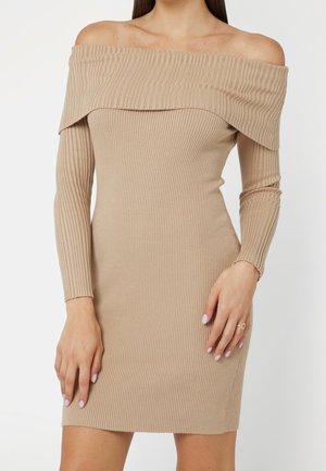 Femme portant une robe beige en maille côtelée, épaules dénudées, manches longues, les mains reposant sur les côtés et un vernis à ongles rose pâle.