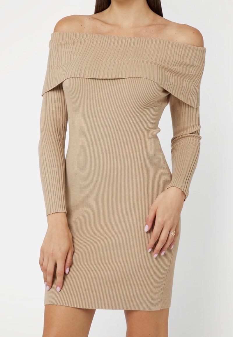 Femme portant une robe beige en maille côtelée, épaules dénudées, manches longues, les mains reposant sur les côtés et un vernis à ongles rose pâle.