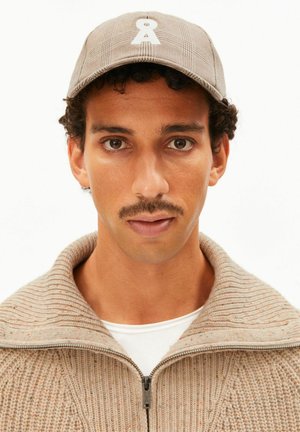 Man with mustache wearing beige plaid cap with white symbol and beige knit zip-up sweater over white shirt, looking forward.