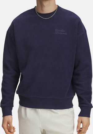 Homme portant un sweat-shirt Under Armour bleu foncé et une chaîne en argent, associé à un pantalon clair, debout devant un fond blanc.