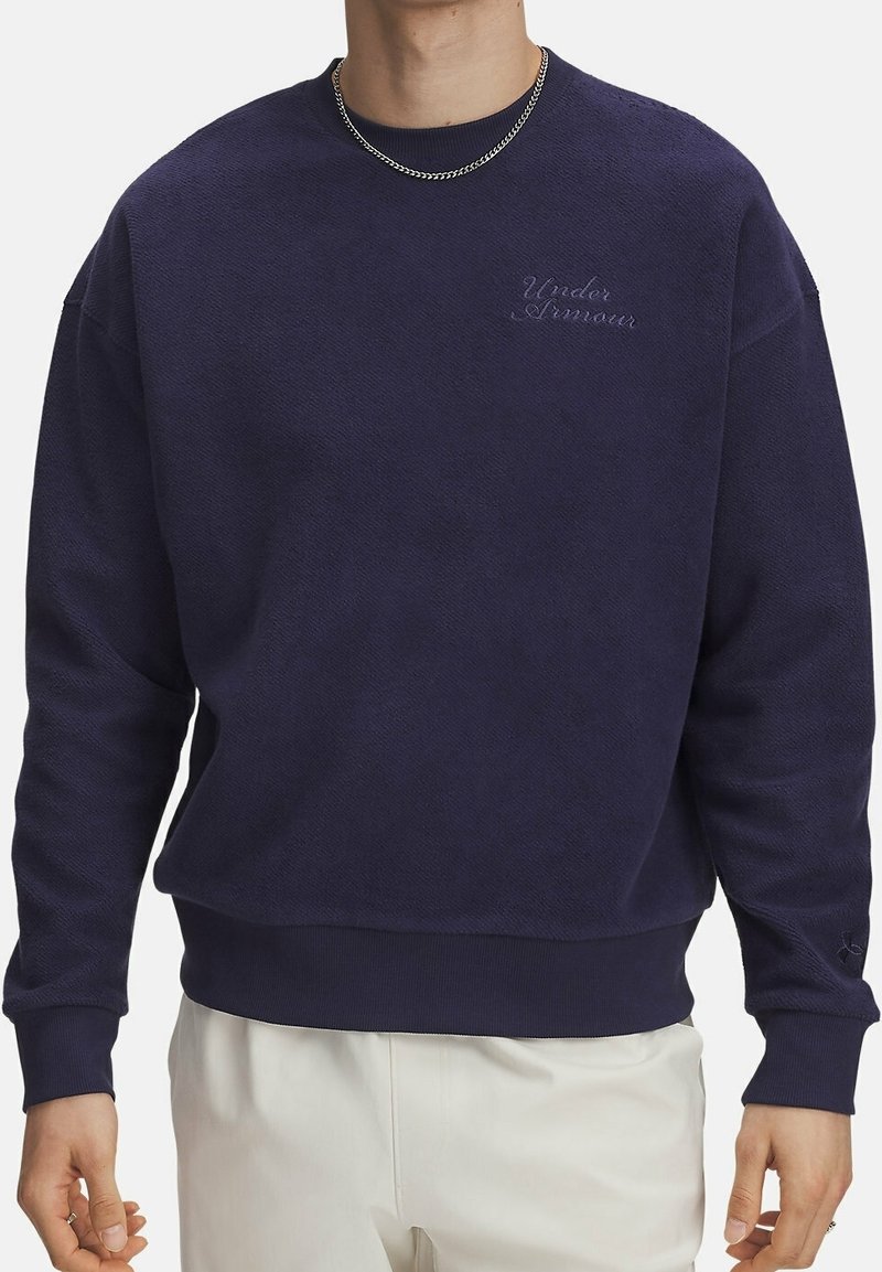 Homme portant un sweat-shirt Under Armour bleu foncé et une chaîne en argent, associé à un pantalon clair, debout devant un fond blanc.