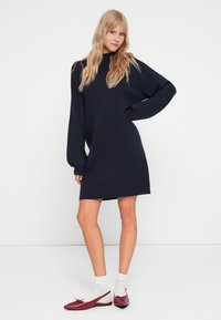 Μπλε φόρεμα φόρμα με ψηλό λαιμό, μακριά μανίκια και χαλαρή εφαρμογή, σε συνδυασμό με κόκκινα σαμπό και λευκές κάλτσες μέχρι τον αστράγαλο.