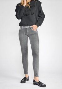 Žena v šedých úzkých džínách, černé mikině s textem a černých mokasínech, stojí s jednou rukou v kapse na jednobarevném pozadí.