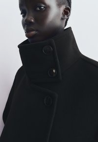 Svart ullrock med hög krage, som har tre stora svarta knappar och en slät textur, visad mot en ljus bakgrund.