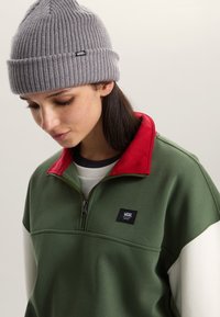 Zelená mikina na zip s červeným límcem, bílými detaily a malým černým logem Vans. Na hlavě šedá pletená čepice s žebrovaným vzorem.