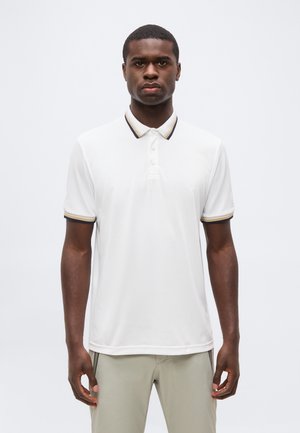 Mężczyzna w białej koszulce polo z krótkim rękawem z czarnym i beżowym wykończeniem na kołnierzu i mankietach, stojący na jednolitym tle.