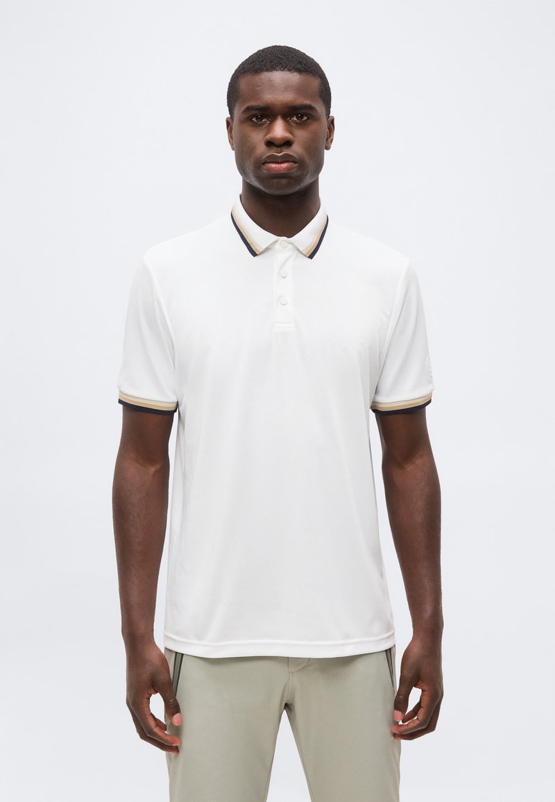 Άνδρας που φοράει λευκό πόλο μπλουζάκι με κοντά μανίκια και μαύρη και μπεζ επένδυση στο γιακά και στα μανίκια, στέκεται σε μονόχρωμο φόντο.