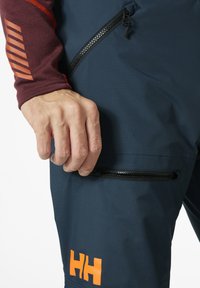 Marineblauwe waterdichte broek heeft een zwarte ritszak en een oranje "HH" logo. Het materiaal heeft een gladde, gestructureerde afwerking.