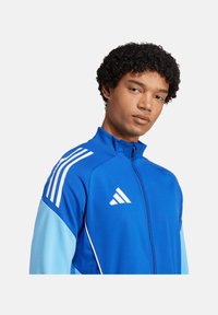 Blaues Zip-Jackett mit hellblauen Ärmeln, weißen Akzenten und drei weißen Streifen auf den Schultern. Auf der Brust befindet sich ein adidas-Logo.
