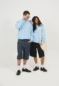 Lyseblå hettegensere med hvit logo, mørk denimshorts, svarte shorts, hvite sokker og svarte sko; modellen holder en flettet bag.