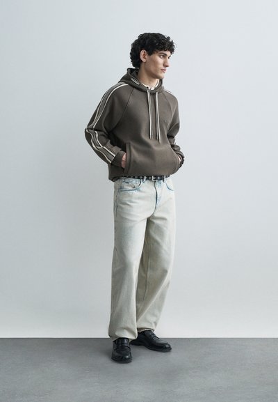 Καφέ φούτερ με κορδόνια και ρίγες στο πλάι, σε συνδυασμό με ανοιχτό μπλε τζιν παντελόνι με φαρδιά μπατζάκια και μαύρα παπούτσια, στέκεται μπροστά σε ουδέτερο φόντο.