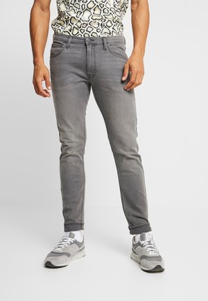 Man met strakke grijze spijkerbroek, ingestopt shirt met luipaardprint, witte sokken en grijs-witte sneakers, staand tegen een effen achtergrond.