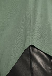 Zelený lehký textilní top se špičatým lemem, vrstvený přes černý texturovaný materiál, ukazující kontrastní barvy a látky.