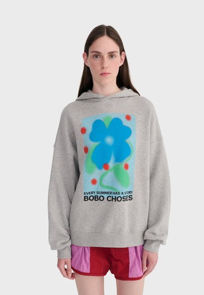 Femme portant un sweat à capuche gris avec une grande fleur bleue et le texte "Chaque été a une histoire BOBO CHOSES", assorti à un short rouge et rose.