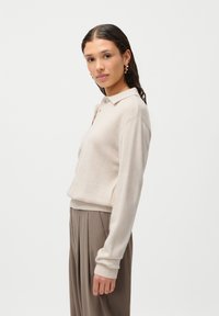 Suéter bege de malha com colarinho estilo polo, punhos e barra canelados, combinado com calças pregueadas em tom castanho claro. Textura suave, corte descontraído.