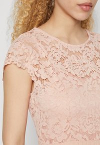 Robe en dentelle rose avec des manches courtes à bords festonnés et un motif floral. Le tissu semble texturé, mettant en valeur des détails de dentelle complexes.
