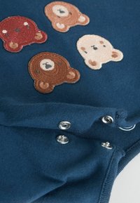Marinblå tygbodysuit med fyra broderade björnskallar i brun, röd och krämfärg. Tryckknappar i botten för enkel åtkomst.