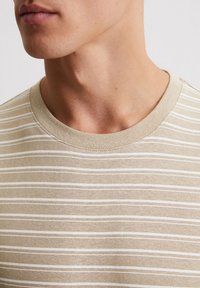 T-shirt beige à rayures avec un col rond, présentant de fines rayures horizontales blanches. Texture douce avec des détails de couture visibles.