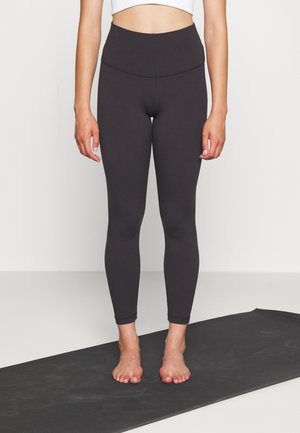 Leggings negros de cintura alta con una textura suave, diseño ajustado y largo hasta el tobillo, combinados con un top corto blanco, sobre un tapete de yoga negro.