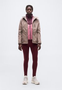 Άτομο που στέκεται, φοράει μπεζ μπουφάν, ροζ φλις με φερμουάρ, μπορντό κολάν και μπεζ ορειβατικά μποτάκια, με φόντο έναν απλό τοίχο.