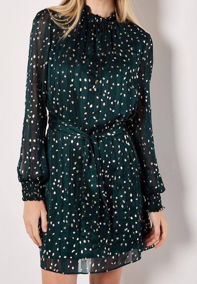 Femme portant une robe vert foncé à manches longues avec des pois dorés métalliques, un col montant à volants et une ceinture à nouer à la taille.