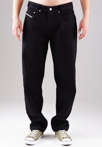 Schwarze Jeans aus Denim mit geradem Schnitt, ausgestattet mit kupferfarbenen Nieten, fünf Taschen und einer dezenten, verblassten Textur; kombiniert mit grünen Sneakers.