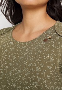 Oliwkowy bawełniany top z kwiatowym wzorem w kolorze off-white. Posiada okrągły dekolt oraz ozdobne metalowe akcenty na ramionach.
