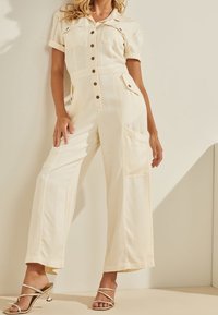 Krémový overal s krátkými ohrnutými rukávy, předním zapínáním na knoflíky a několika kapsami; vyrobený z hladkého materiálu s volným střihem.