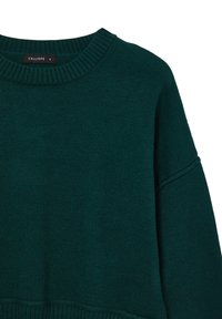 Pull en maille vert foncé avec un col rond côtelé et des poignets, présentant une coupe légèrement ample et une texture lisse. Étiquette de marque visible.