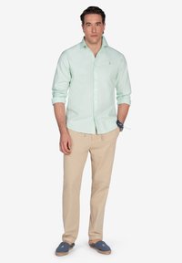 Camisa verde claro de botones hecha de algodón, combinada con pantalones beige y alpargatas azules. La camisa presenta un cuello y un sutil detalle de logotipo.
