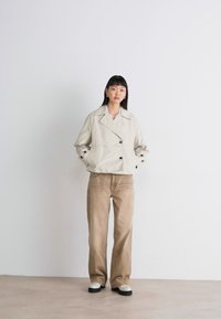 Beige lyhyt jakku, jossa on kaksinkertaiset napit, yhdistettynä vaaleanruskeisiin housuihin ja valkoisiin kenkiin. Sileä kangas ja minimalistinen muotoilu.