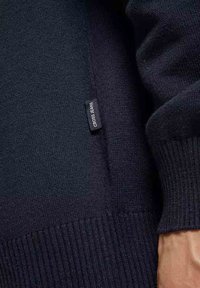 Sweter w ciemnoniebieskim kolorze, wykonany na drutach, z prążkowanymi mankietami i ściągaczem u dołu, z małą metką "Cross Jeans" przyszytą na bocznym szwie.