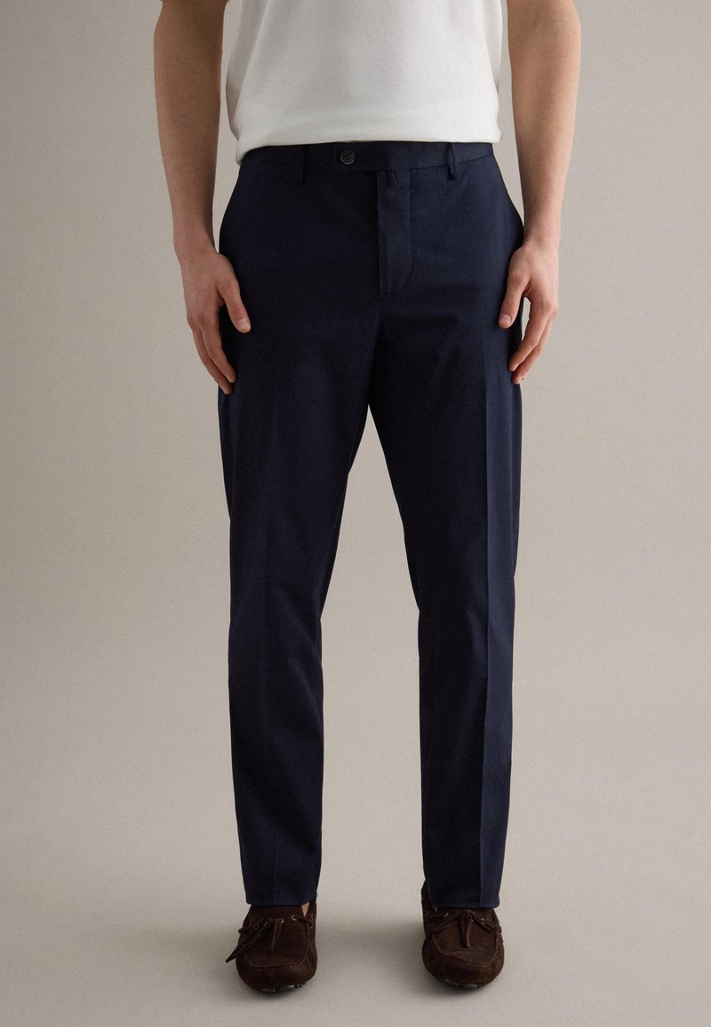 Mann trägt marineblaue maßgeschneiderte Hose und braune Wildleder-Loafer, steht vor einem einfarbigen beigen Hintergrund.