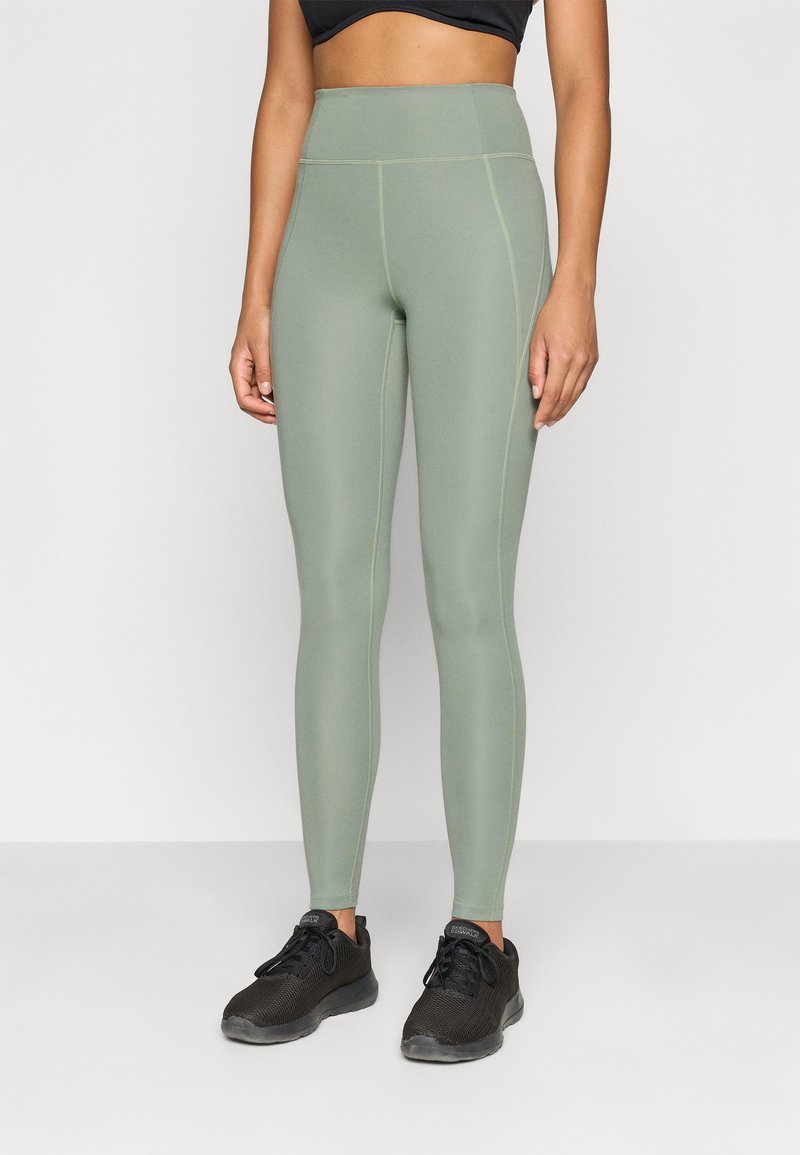 Leggings de tiro alto en verde claro con costuras amarillas en contraste, fabricados en un tejido elástico, combinados con zapatillas deportivas de malla negras.