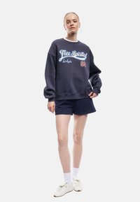 Marineblauwe sweatshirt met een ronde hals en een losse pasvorm, met een witte en rode afbeelding "Free Spirited Los Angeles 93". Gecombineerd met donkere shorts en witte sneakers.