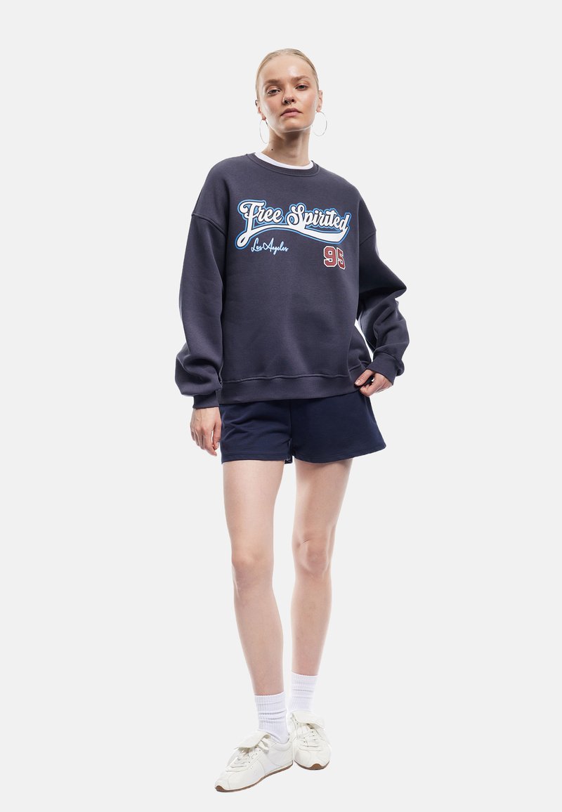 Marineblauwe sweatshirt met een ronde hals en een losse pasvorm, met een witte en rode afbeelding "Free Spirited Los Angeles 93". Gecombineerd met donkere shorts en witte sneakers.