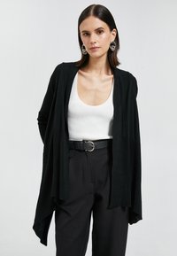 Cardigan de punto negro con un diseño abierto, que presenta un ajuste relajado. Se lleva sobre una camiseta blanca de escote redondo y se combina con pantalones negros de tiro alto.