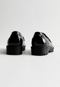 Ett par svarta glänsande läderloafers med kraftiga sulor sedda bakifrån på en enkel vit yta.