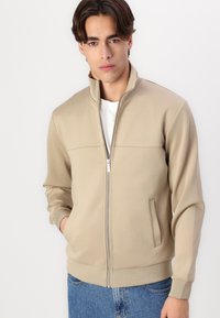 Beige Zip-Jacke mit hohem Kragen, zwei Seitentaschen und dezenten Nähten. Getragen über einem weißen T-Shirt, kombiniert mit blauen Jeans.